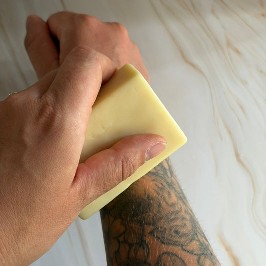 A close-up of a person's tattooed arm and hand holding a pale yellow rectangular bar of moisturiser against their forearm, with a light wood plank background.
