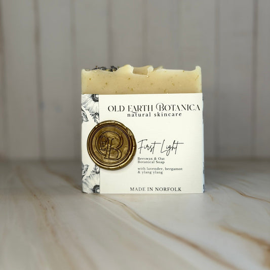 A bar of Old Earth Botanica natural skincare soap labeled 'First Light' resting on a light marble surface against a white wood-panelled background.