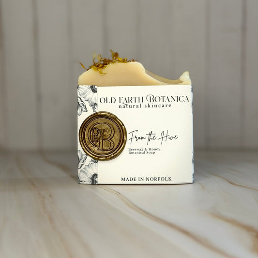 A bar of Old Earth Botanica natural skincare soap, wrapped in a white paper sleeve with black botanical illustrations and a gold wax seal, sitting on a light marble surface against a gray wood-paneled background.