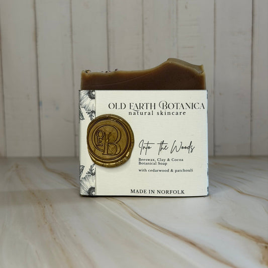 A bar of Old Earth Botanica 'Into the Woods' botanical soap, wrapped in a cream-colored paper sleeve with a gold wax seal and black text, set against a light wood paneled background.