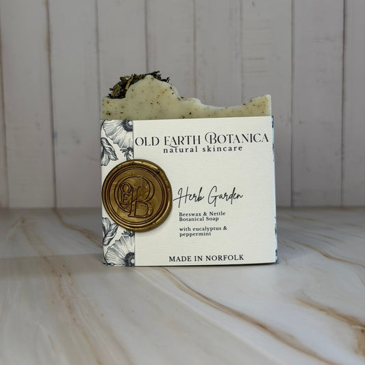A bar of Old Earth Botanica Herb Garden soap, wrapped in a white paper sleeve with a gold wax seal, set against a white wood paneled background.