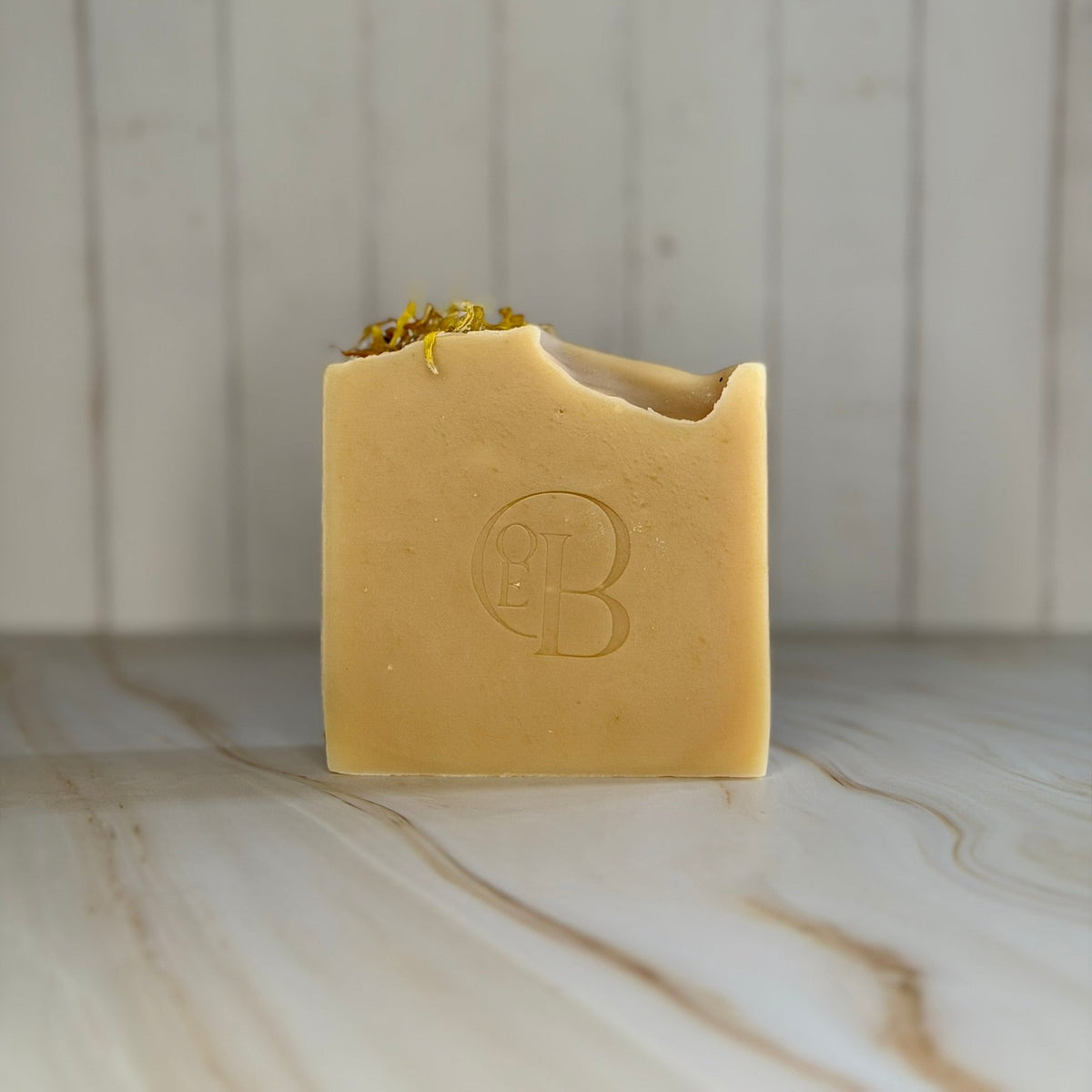 A square bar of pale yellow soap and dried botanicals on the edge, standing on a light marble surface against a white wood plank background. The soap is embossed with a circular 'OEB' logo.