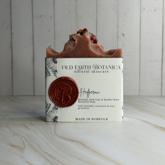 A handmade bar of soap from Old Earth Botanica, wrapped in a white paper sleeve with a berry red wax seal, sitting on a light marble surface against a white wood paneled background.