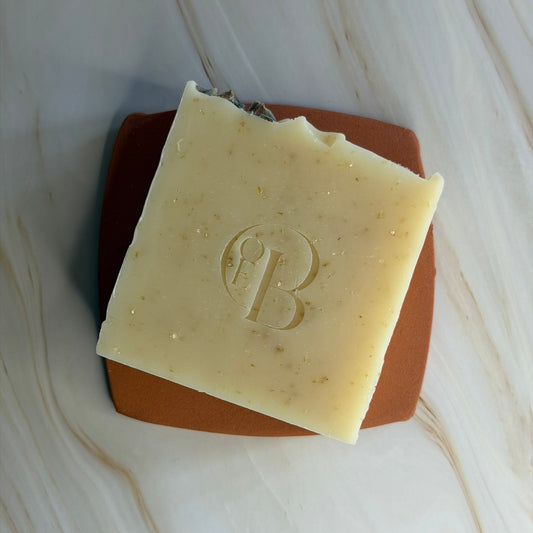 A square bar of pale yellow soap with a raised monogram logo, resting on a terracotta-coloured square tile over a white marble surface.