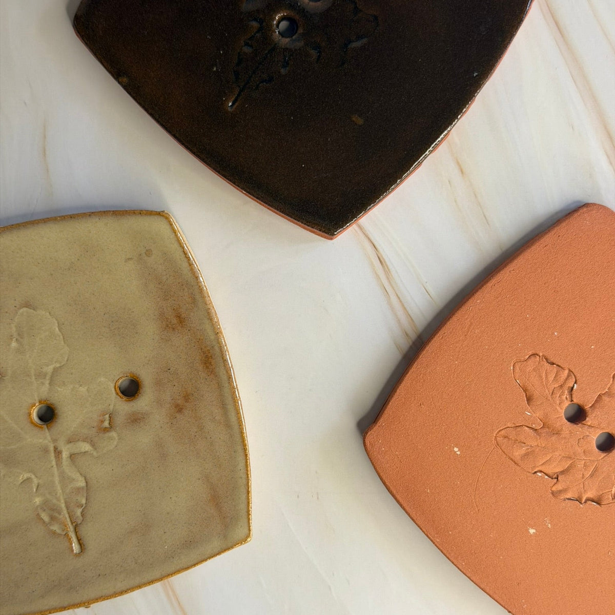 Three ceramic soap dishes with a leaf motif and three holes, shown in dark brown, beige, and terracotta orange on a light wood background.