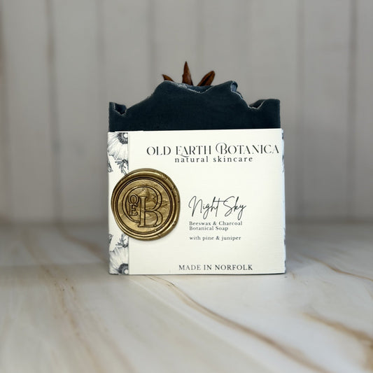 A bar of Old Earth Botanica natural skincare soap labeled 'Night Sky' with a white paper wrapper and a gold wax seal, standing on a light marble surface against a white wood-paneled background.