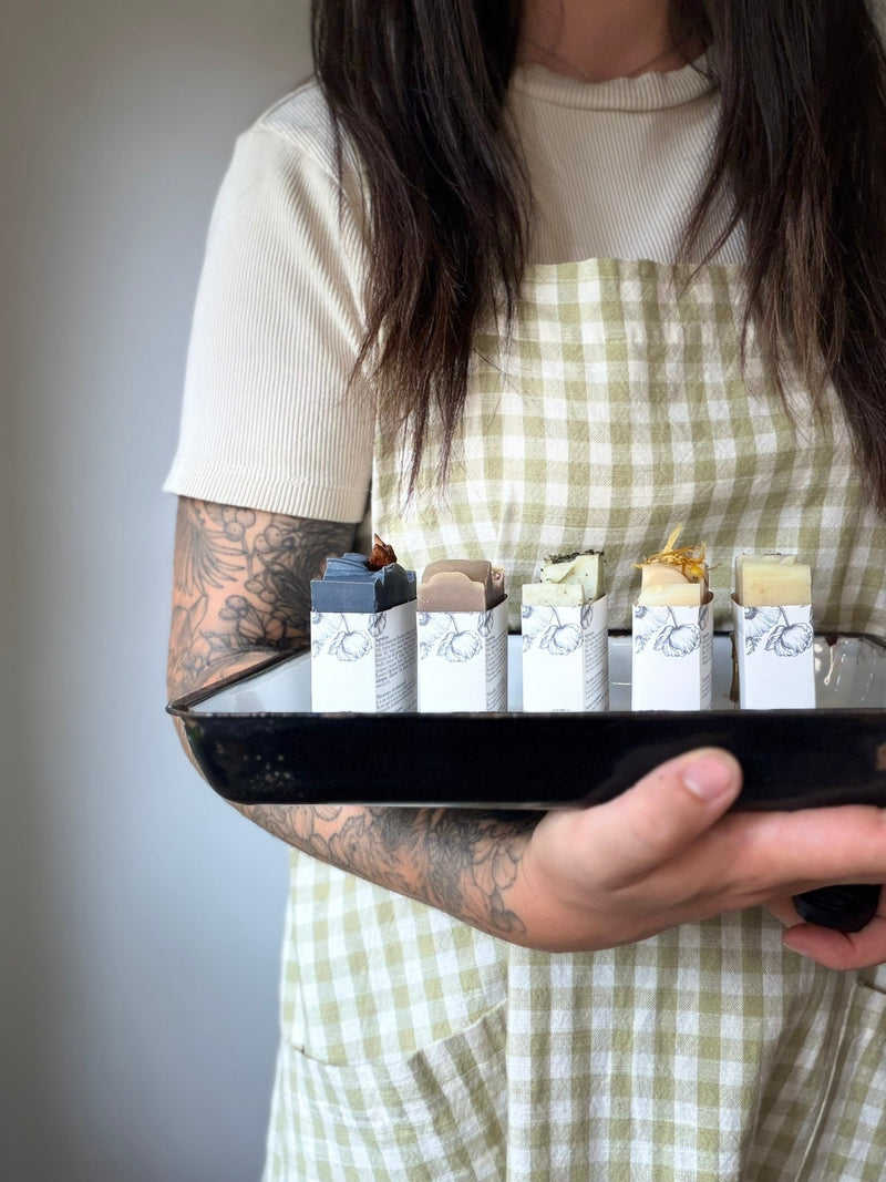 A woman with dark hair and a pale green gingham apron with a botanical sleeve tattoo holding an old enamel tray with 5 packaged soaps.