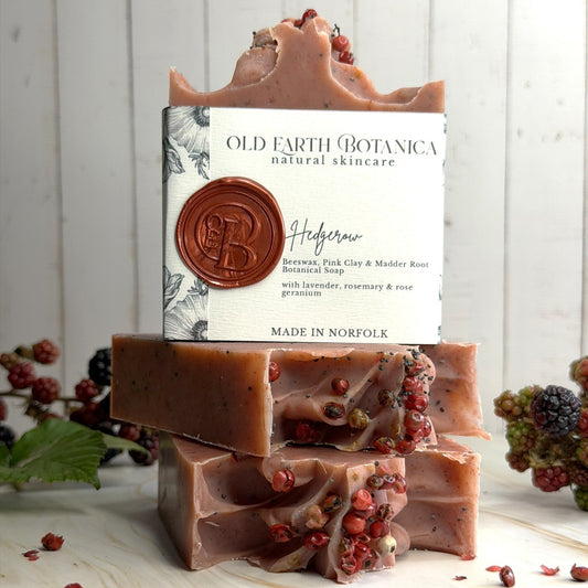 Three stacked bars of Old Earth Botanica Hedgerow Soap on a light wood surface with a white wood plank background. The soap is a dusty mauve-brown color with visible dried berries and the top bar has its paper label facing forward.