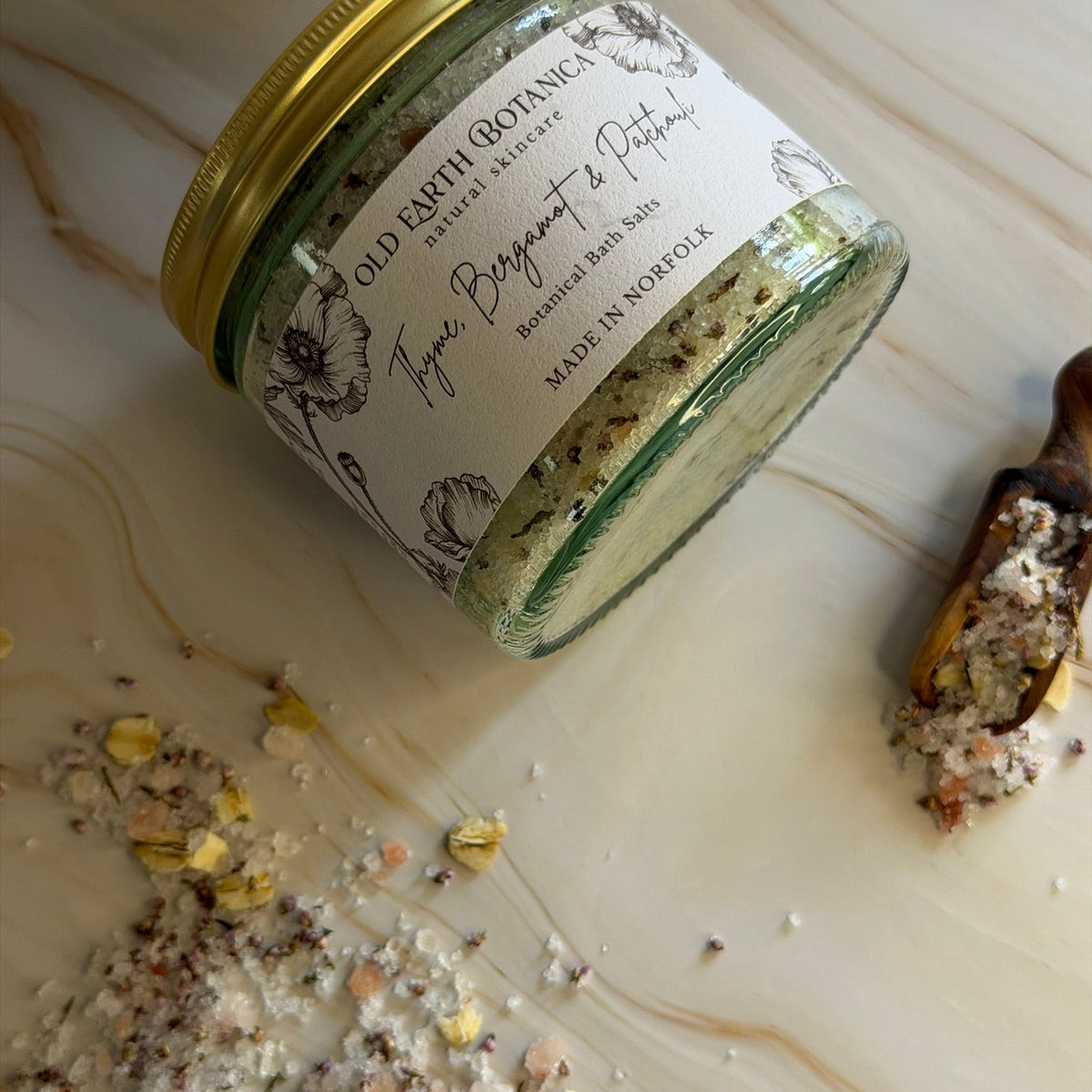 A glass jar of Old Earth Botanicas Thyme, Bergamot & Patchouli Botanical Bath Salts with a gold lid and textured label, shown on a marble surface with some salt spilled and a wooden scoop nearby.