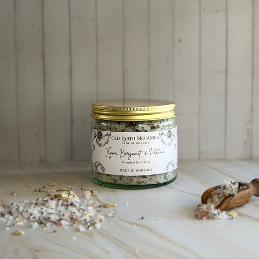 A glass jar of Old Earth Botanica Thyme, Bergamot & Patchouli Botanical Bath Salts in a clear glass jar with a gold aluminium lid and textured label, styled on a marble surface with scattered bath salts and a wooden spoon against a light wood panelled background.
