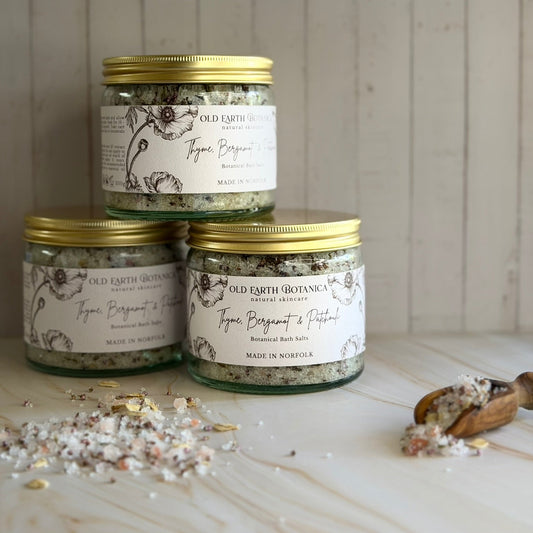 Three glass jars of Old Earth Botanicas Thyme, Bergamot & Patchouli Botanical Bath Salts are stacked in a pyramid on a light marble surface with scattered salt crystals and a wooden spoon in the foreground, set against a light wood panelled background.