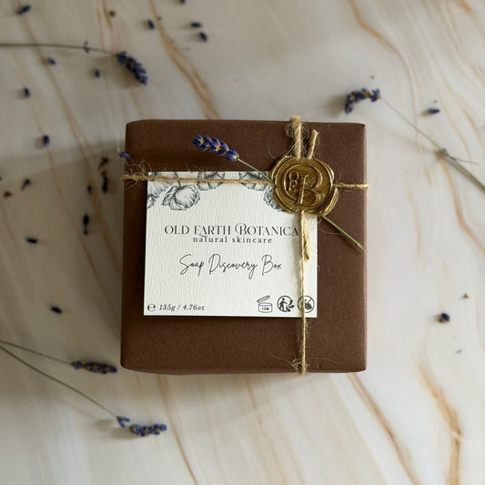 A brown gift box wrapped in twine and a cream paper label reading 'Old Earth Botanicas Soap Discovery Box,' tied with a gold seal, placed on a light marble surface with scattered dried lavender sprigs.