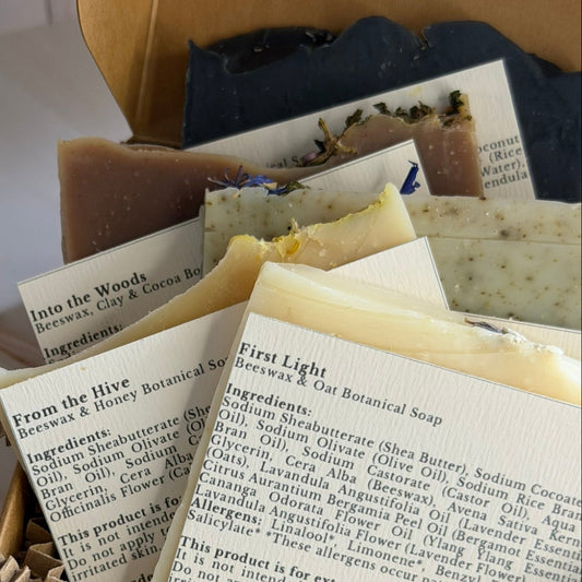 A cardboard gift box filled with handmade artisan soaps and printed paper inserts, photographed on a light wood surface with a white tiled background.