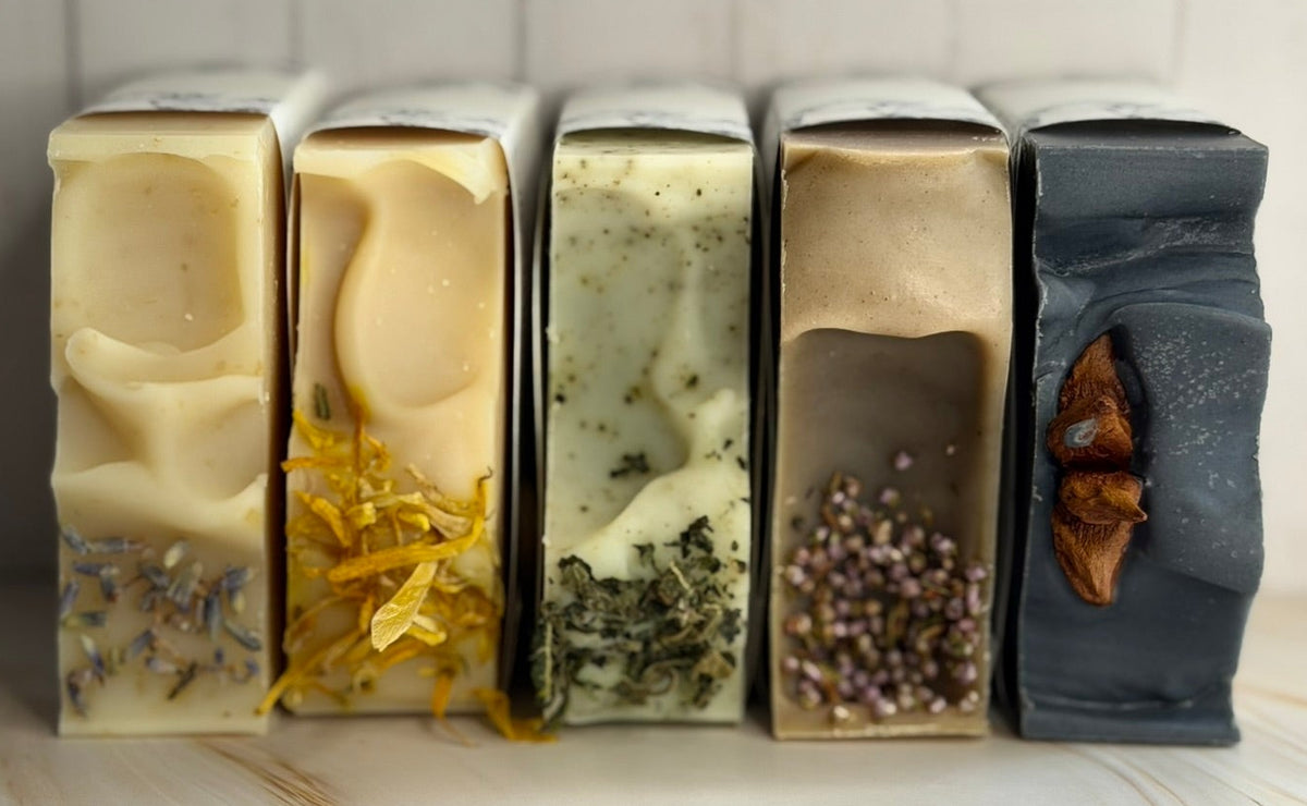 Five artisanal handmade soap bars with embedded botanicals and spices, arranged in a row on a light marble surface against a white wood-paneled background.