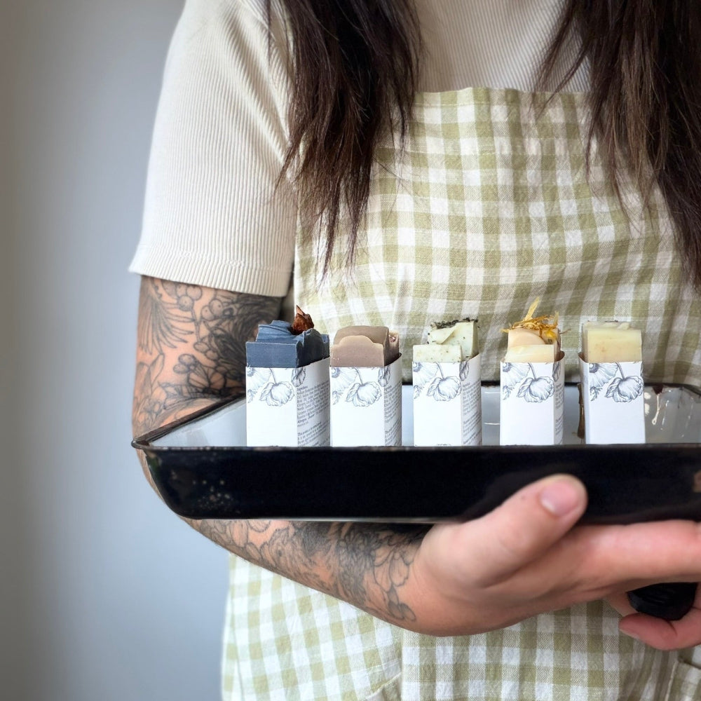A person wearing a cream ribbed top and a light green and white gingham apron holds a black tray with five small white boxes of what appear to be soaps or similar bar products. The background is a plain light gray wall.