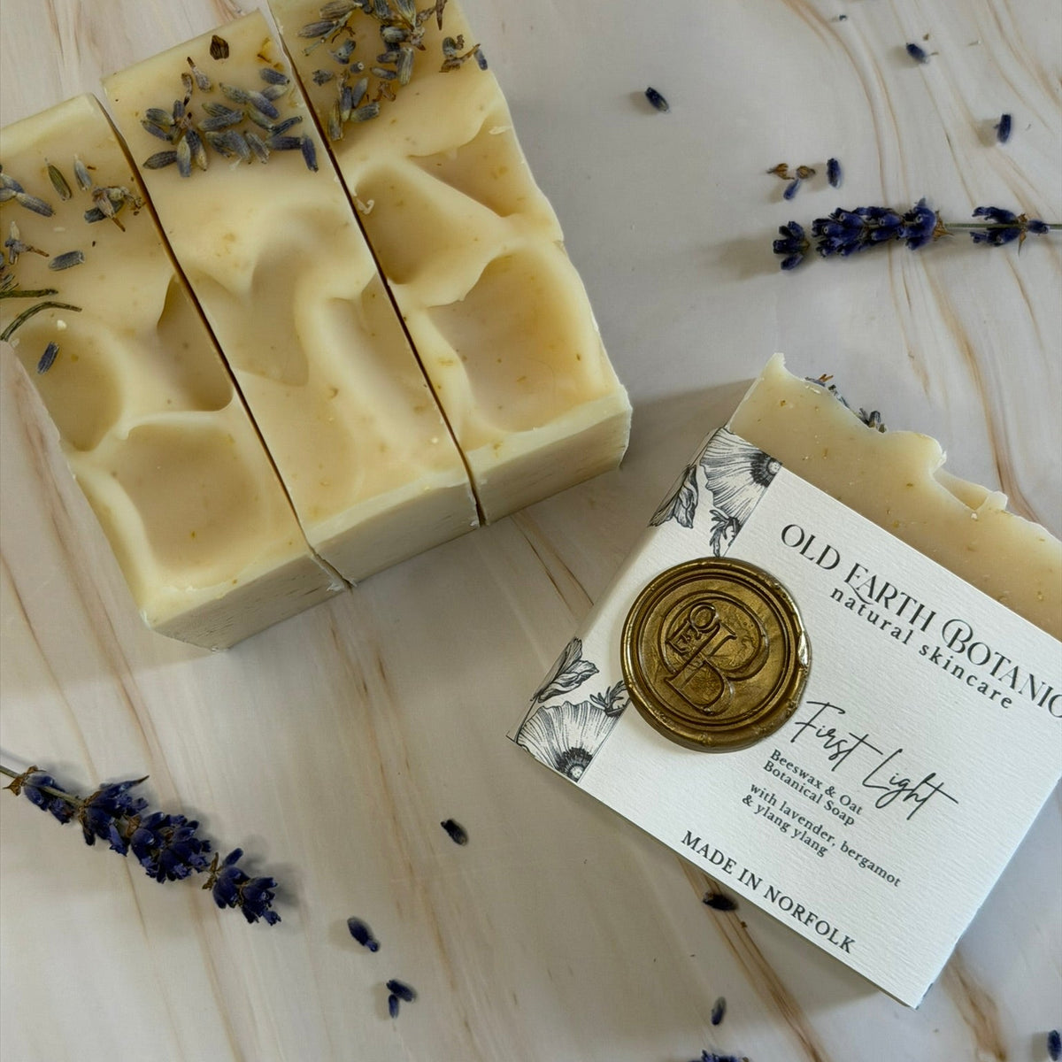 Three bars of Old Earth Botanica natural skincare soap, 'First Light' in, displayed on a light marble background with scattered dried lavender sprigs. One bar is sliced to show the texture, and another is resting on its white paper packaging card with a gold wax seal.