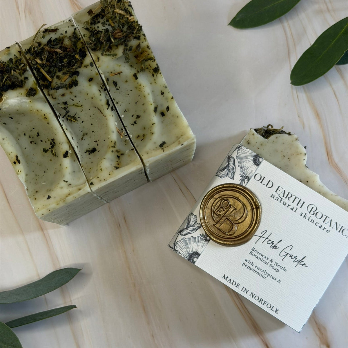 Two bars of handmade soap on a light marble surface with green leaves. One bar is sliced to show the textured, speckled cream and green herbal soap, and the other is wrapped in a white paper sleeve with a gold wax seal reading 'OLD EARTH BOTANICA Herb Garden Soap'.