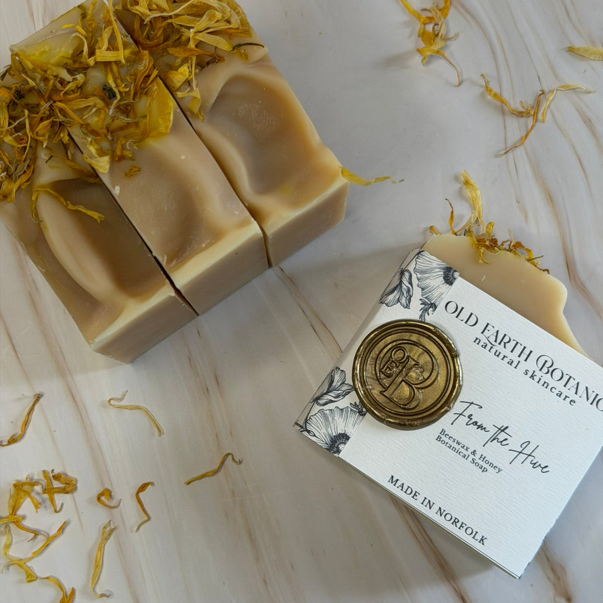 A bar of handmade soap in a cream-beige color, topped with dried yellow botanical petals, shown next to its white paper packaging box with a gold wax seal on a light marble surface.