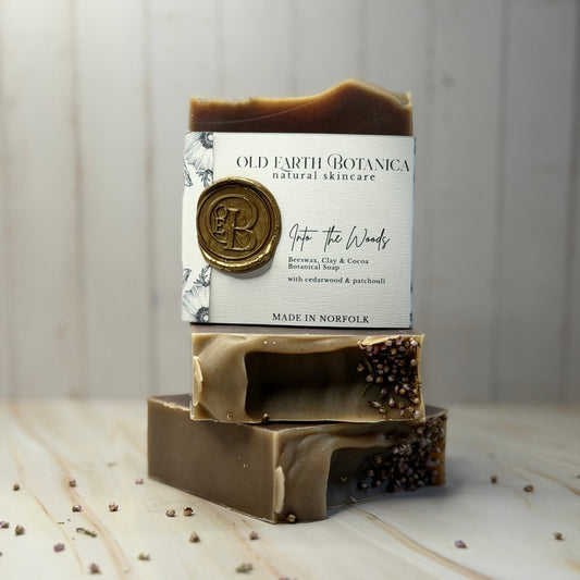 Three stacked bars of Old Earth Botanica handmade soap on a light wood surface with a white wood plank background. The top bar is wrapped in a white paper label with a gold wax seal, and the soap bars show marbled brown and beige patterns with dark speckles.
