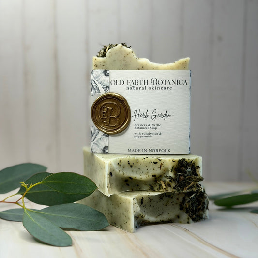 Two stacked bars of Old Earth Botanica Herb Garden natural skincare soap, featuring a white label with a gold wax seal, displayed on a light wooden surface with eucalyptus leaves.