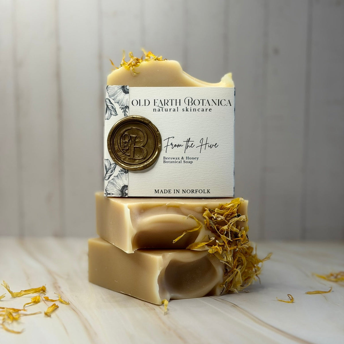 A stack of three handmade natural soap bars with dried yellow botanicals, featuring a white paper label on the top bar that reads 'OLD EARTH BOTANICA natural skincare' and 'From the Hive'.