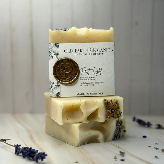 Two stacked bars of Old Earth Botanica First Light botanical soap, featuring a cream-colored bar on top with a white paper label and a gold wax seal, and a natural off-white bar below with visible dried lavender and herbs. The soap is styled on a light wood surface with scattered lavender sprigs against a white wood-paneled background.