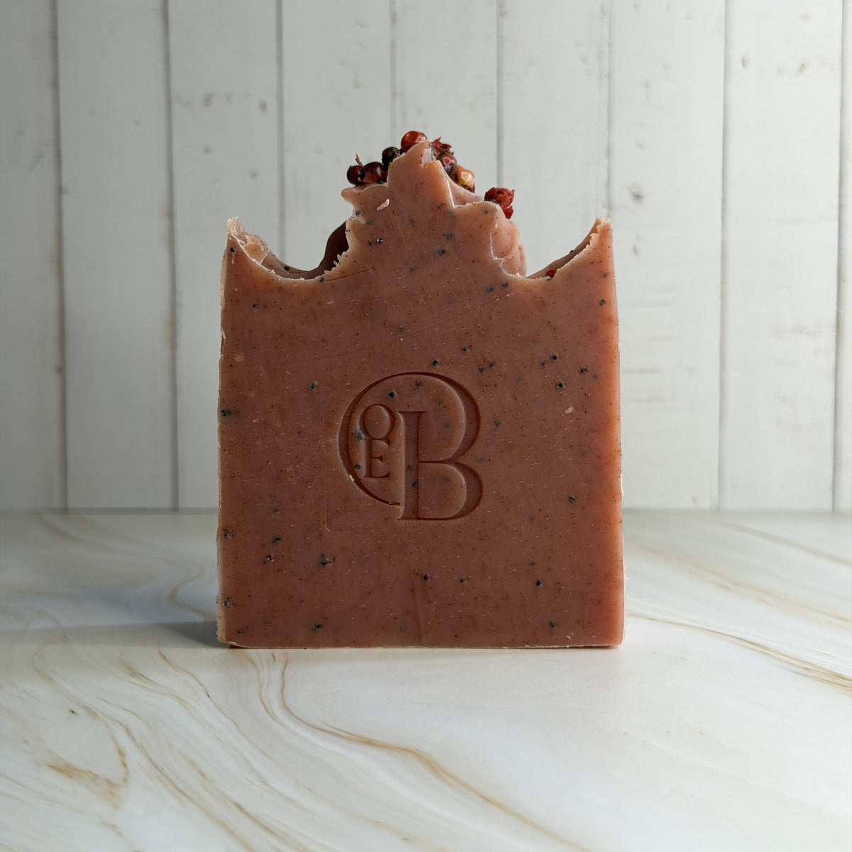 A rectangular bar of reddish-brown soap with dark speckles and a decorative top, standing upright on a light marble surface against a white wood plank background. The soap is embossed with a circular monogram logo.