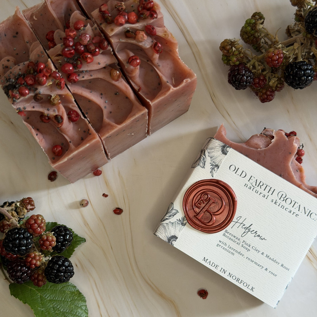A styled flat-lay of a handmade soap bar and its branded paper packaging on a light wood surface. The soap is a dusty mauve-pink color with dark berry specks and red berries on top.
