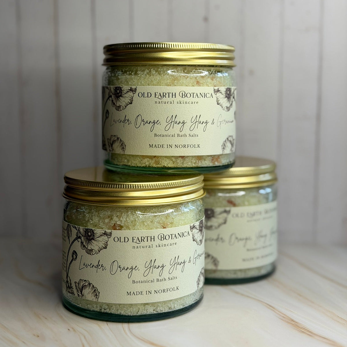 Three glass jars of Old Earth Botanica bath salts with gold lids, arranged on a light wood surface against a white wood paneled background.