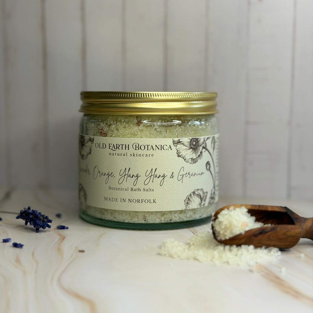 A glass jar of Old Earth Botanica herbal bath salts with a gold lid, styled on a marble surface with a wooden spoon and scattered bath salts against a white wood plank background.