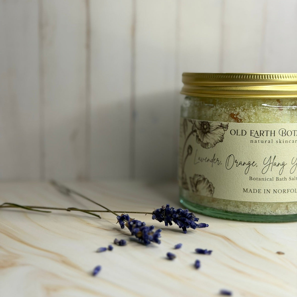 A glass jar of Old Earth Botanicals Lavender, Orange, Geranium and Ylang Ylang botanical bath salts with a gold lid, set on a light wood surface with scattered lavender sprigs against a white wood plank background.