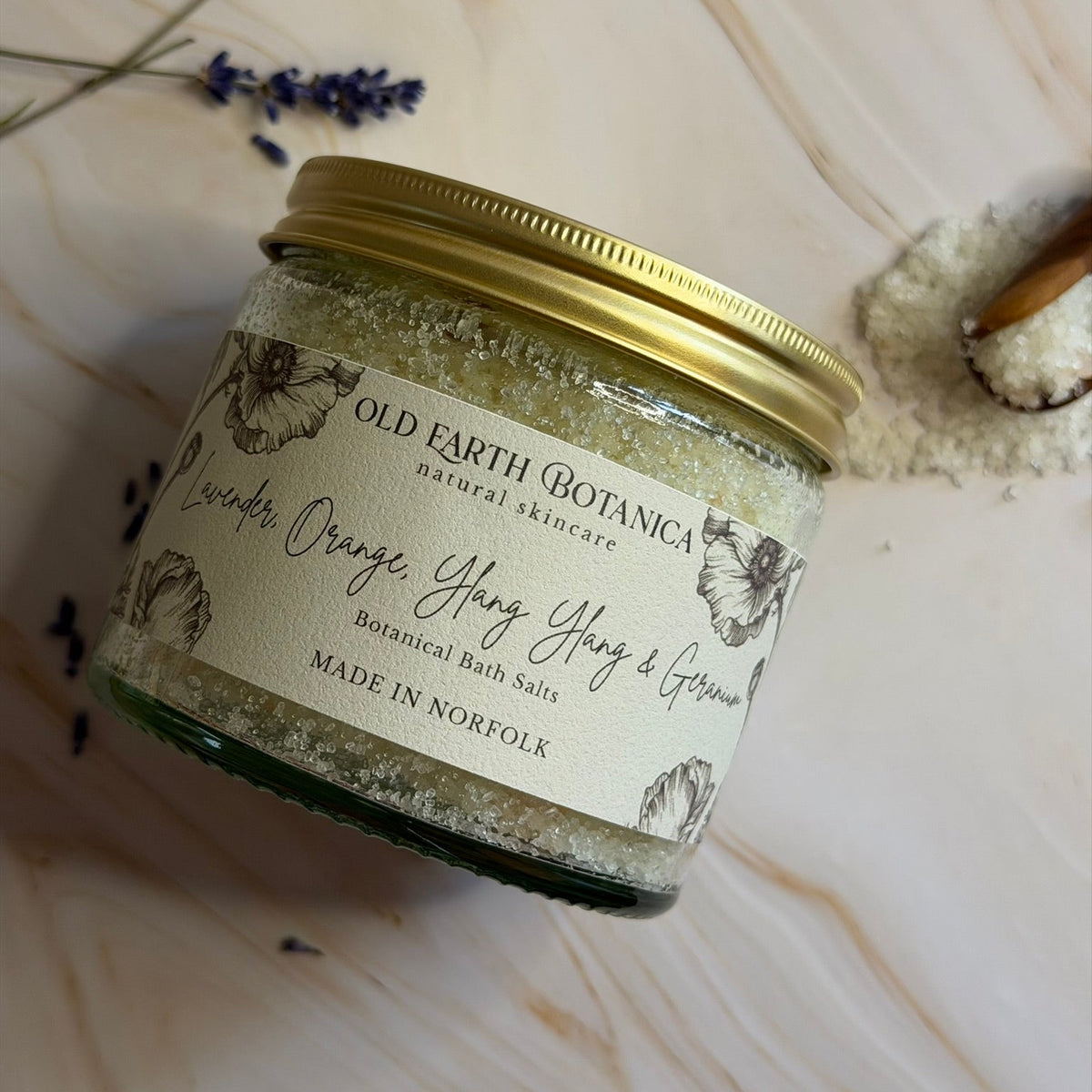 A glass jar of Old Earth Botanica Botanical Bath Salts, featuring a gold metal lid and a cream-colored label with botanical illustrations. The jar is styled on a light beige marble surface with dried lavender sprigs and a small pile of white bath salts nearby.