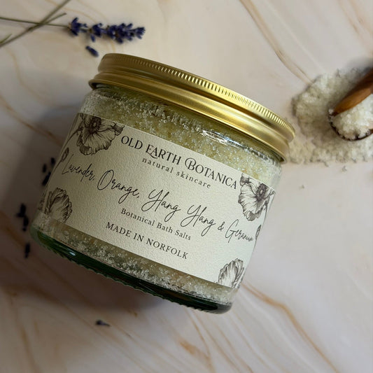A glass jar of Old Earth Botanica Botanical Bath Salts, featuring a gold metal lid and a cream-colored label with botanical illustrations. The jar is styled on a light beige marble surface with dried lavender sprigs and a small pile of white bath salts nearby.