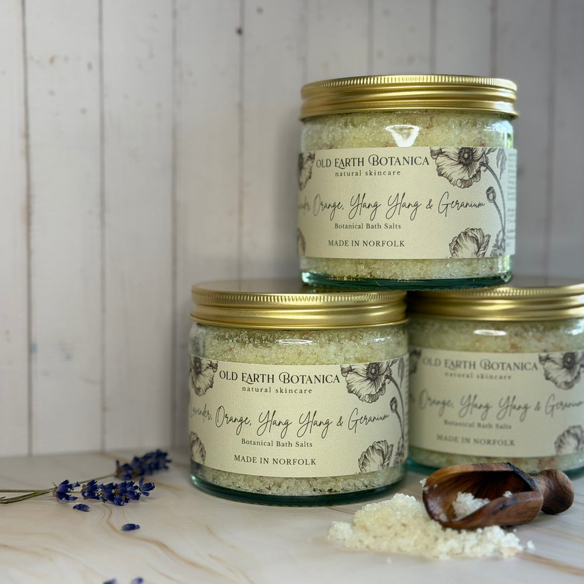 Three glass jars of Old Earth Botanica bath  salts arranged on a light wood surface against a white wood plank background, with lavender sprigs, a wooden spoon, and a small pile of salts nearby.