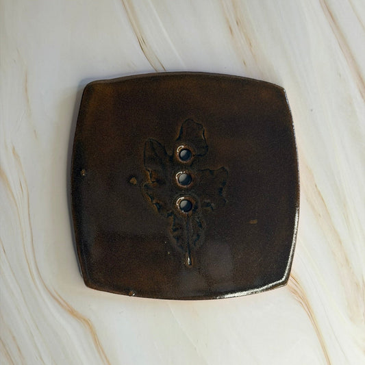 A square dark brown glazed soap tile with rounded corners and a central floral design displayed on a light beige marble surface.