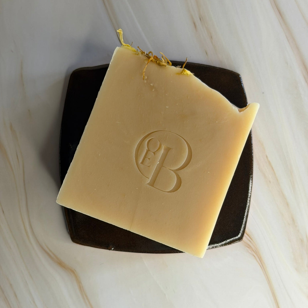A rectangular bar of pale yellow soap with an embossed monogram logo, resting on a dark square dish over a light marble surface.