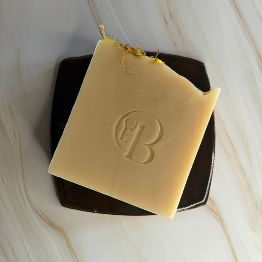 A rectangular bar of pale yellow soap with an embossed monogram logo, resting on a dark square dish over a light marble surface.