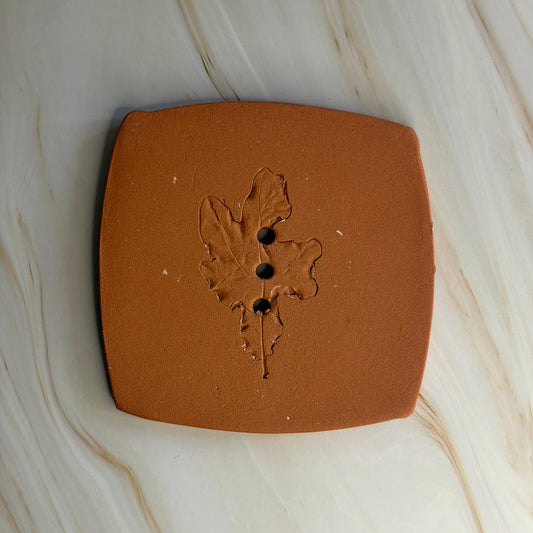 A square terracotta clay coaster with rounded corners and a raised leaf design in the centre, featuring three small holes, shown on a light beige marble surface.