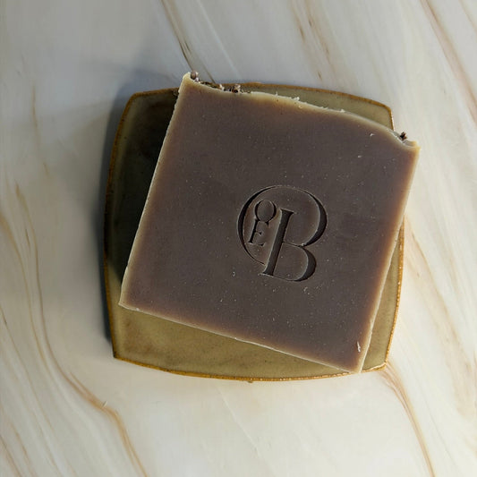 A square bar of soap in a muted taupe-brown color with an embossed monogram logo, resting on a small gold-toned dish over a cream marble surface.