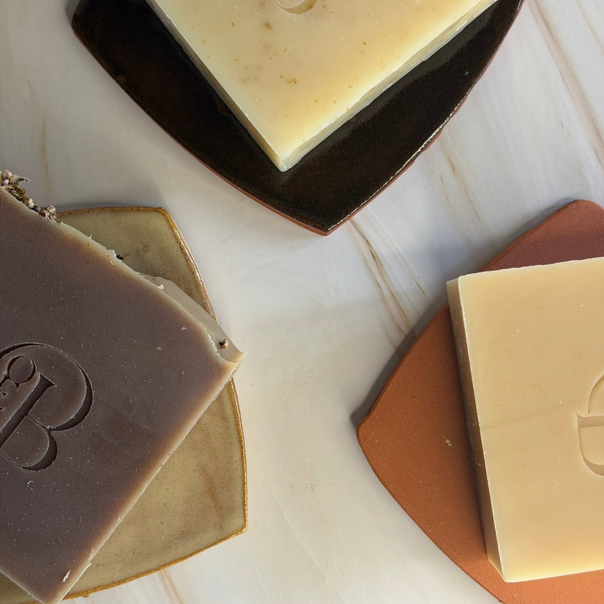 Three artisanal soap bars with embossed 'B' logos, displayed on decorative ceramic coasters on a light marble surface. The soaps are shown in three colors: a creamy beige, a warm tan, and a dark chocolate brown.
