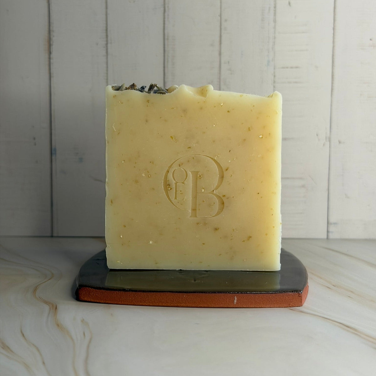 A rectangular bar of pale yellow soap with a textured surface and dried botanicals on top, standing on a dark ceramic and terracotta base against a white wood plank background.