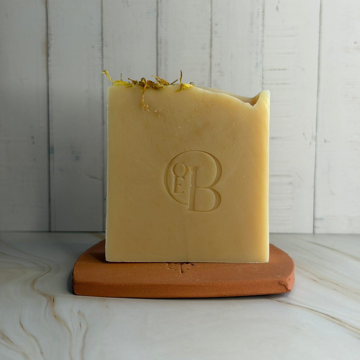 A rectangular bar of pale yellow soap with a rough, rustic edge and dried flower petals on top, standing on a terracotta-colored square base against a white wood plank background.